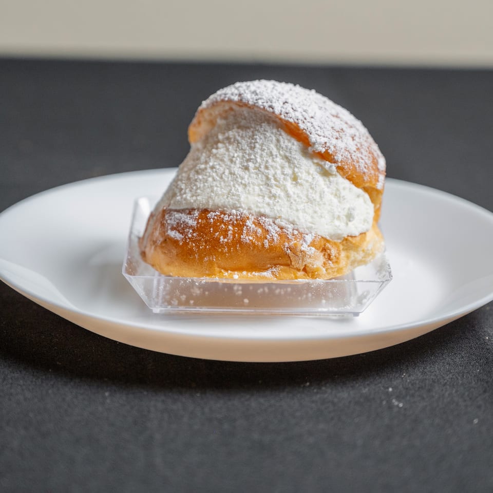 A golden-brown pastry bun, generously filled with white cream and dusted with powdered sugar, on a white plate.
