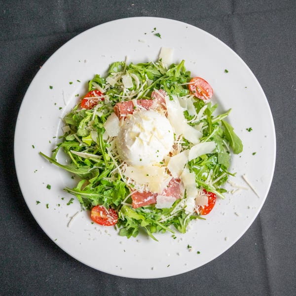 Arugula salad with burrata cheese, prosciutto, cherry tomatoes, and shaved parmesan on a white plate.