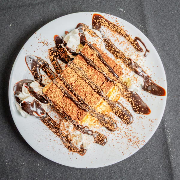 Tiramisu slice with whipped cream, chocolate drizzle, and cocoa powder on a white plate.