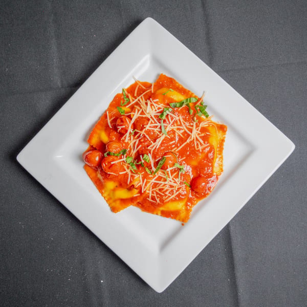 Ravioli with red sauce, cherry tomatoes, shredded cheese, and basil on a white square plate.