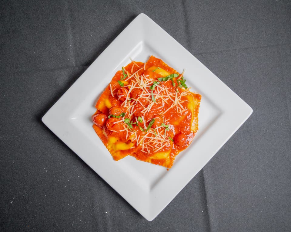 Ravioli with red sauce, cherry tomatoes, shredded cheese, and basil on a white square plate.