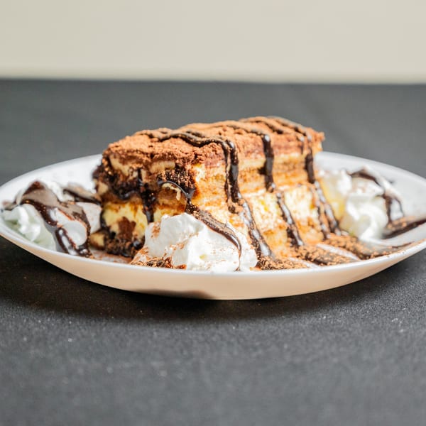 Tiramisu dessert with chocolate drizzle, cocoa powder, and whipped cream on a white plate.