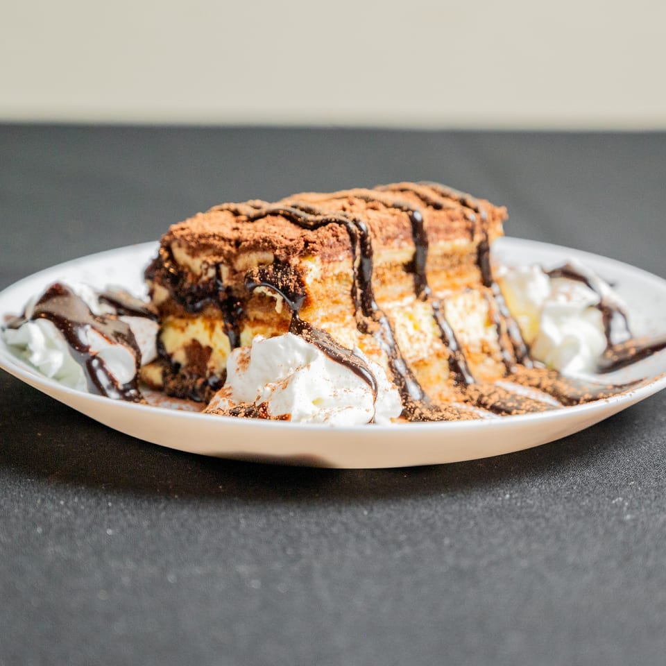 Tiramisu dessert with chocolate drizzle, cocoa powder, and whipped cream on a white plate.
