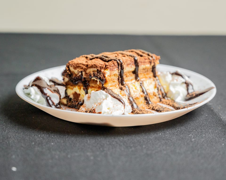Tiramisu dessert with chocolate drizzle, cocoa powder, and whipped cream on a white plate.
