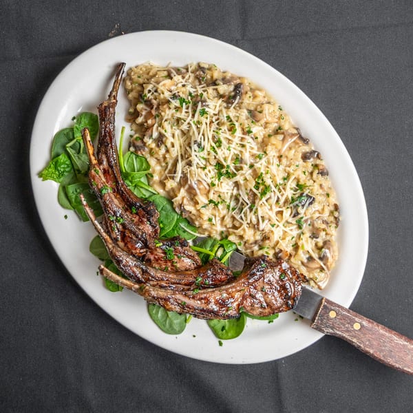 Grilled lamb chops with mushroom risotto, garnished with spinach, cheese, and parsley on a white plate.