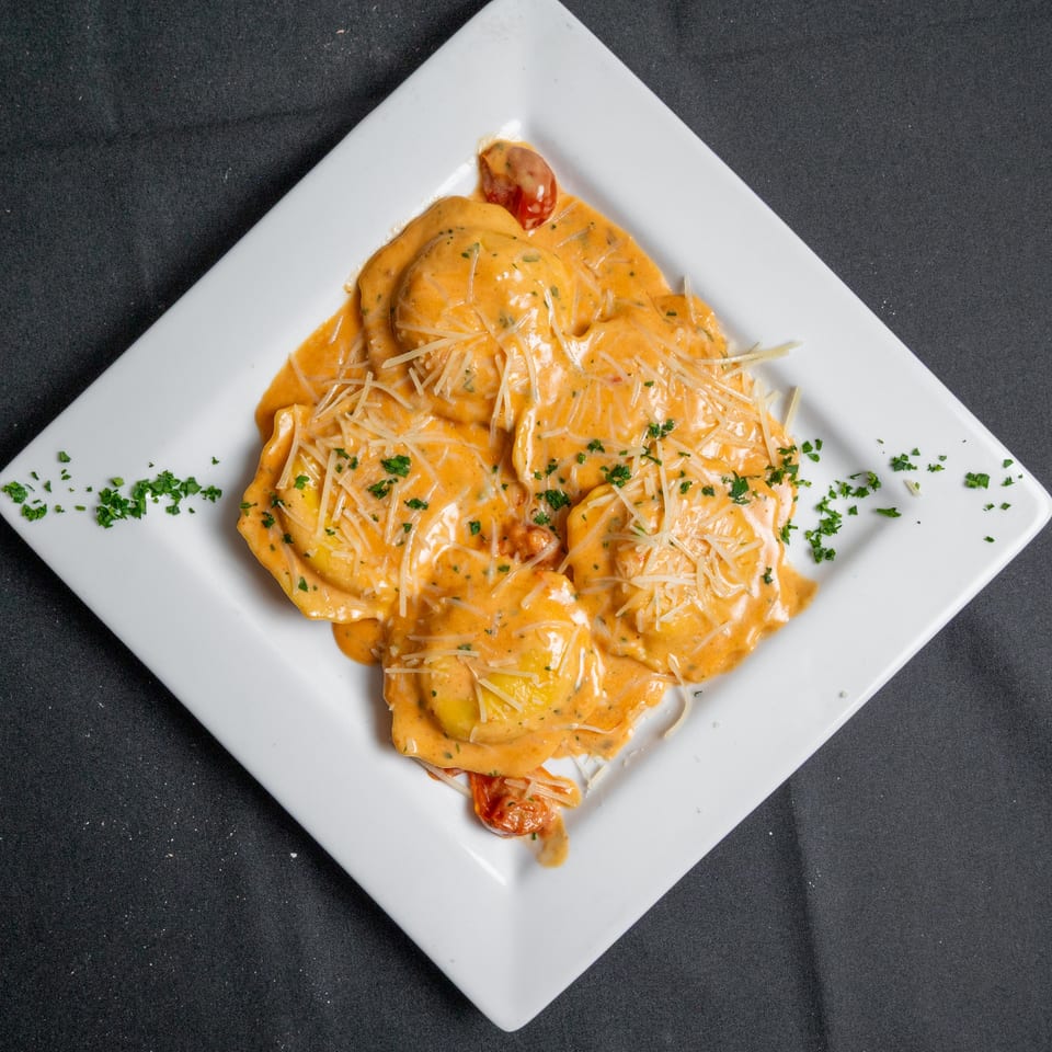 Square white plate of ravioli in a creamy orange sauce, topped with shredded cheese and parsley.