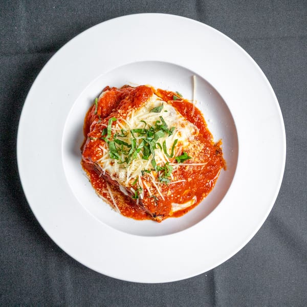 A white bowl holds a serving of lasagna topped with red sauce, melted cheese, and fresh basil.