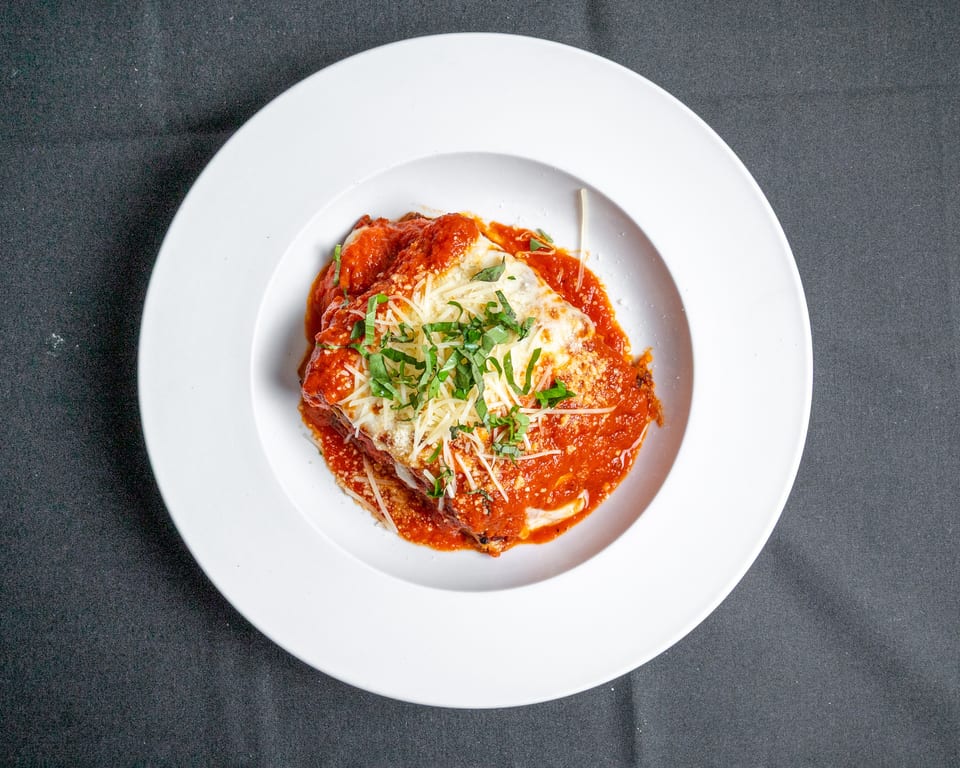 A white bowl holds a serving of lasagna topped with red sauce, melted cheese, and fresh basil.