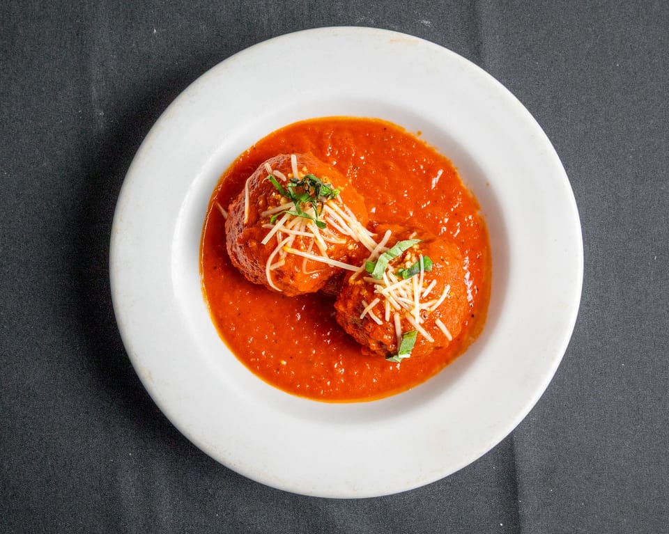Two meatballs in rich red tomato sauce, topped with shredded cheese and fresh basil, in a white bowl.