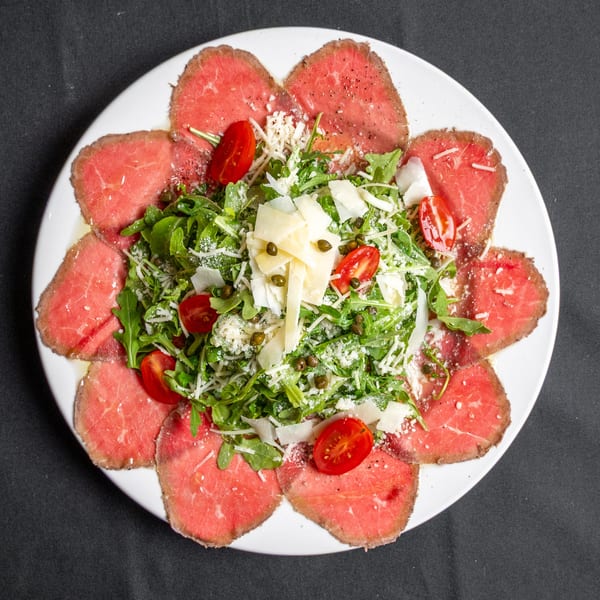 Beef carpaccio with arugula, cherry tomatoes, capers, and parmesan cheese on a white plate.