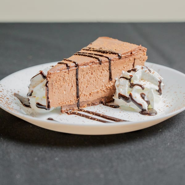 A slice of chocolate cheesecake with whipped cream, chocolate drizzle, and cocoa powder on a white plate.