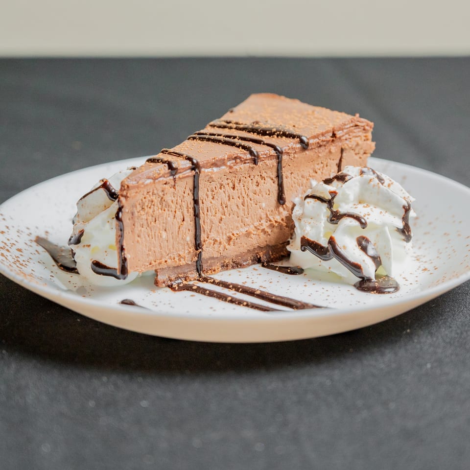 A slice of chocolate cheesecake with whipped cream, chocolate drizzle, and cocoa powder on a white plate.