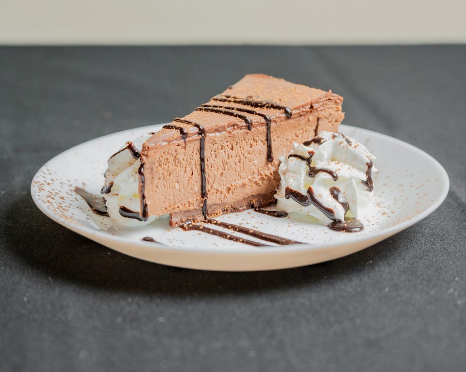 A slice of chocolate cheesecake with whipped cream, chocolate drizzle, and cocoa powder on a white plate.