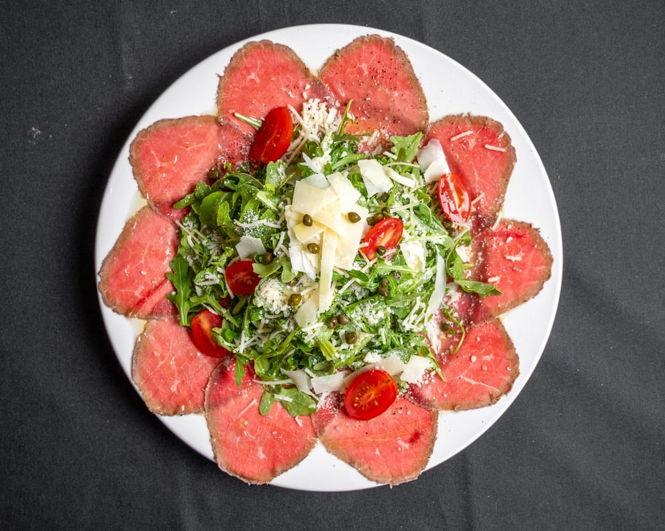 Carpaccio with arugula, cherry tomatoes, capers, and parmesan cheese on a white plate.