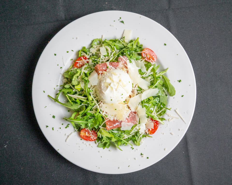 Arugula salad with burrata, prosciutto, cherry tomatoes, and shaved parmesan on a white plate.
