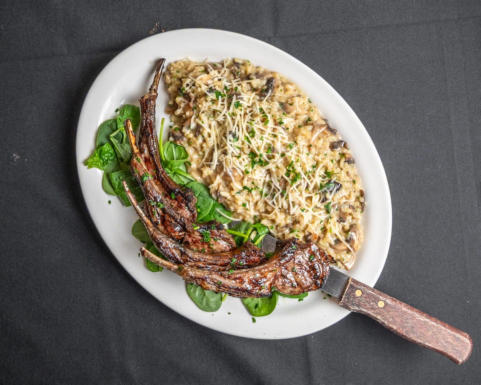 Grilled lamb chops with mushroom risotto, garnished with spinach, cheese, and parsley on a white plate.
