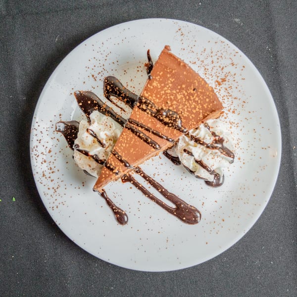 A slice of chocolate cake with whipped cream, chocolate drizzle, and cocoa powder on a white plate.