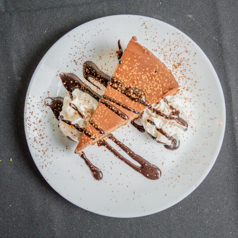 A slice of chocolate cake with whipped cream, chocolate drizzle, and cocoa powder on a white plate.