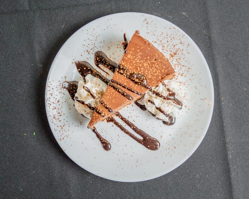 A slice of chocolate cake with whipped cream, chocolate drizzle, and cocoa powder on a white plate.