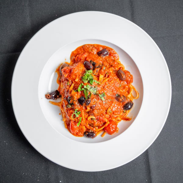 Spaghetti with red tomato sauce, black olives, cherry tomatoes, and fresh herbs in a white bowl.