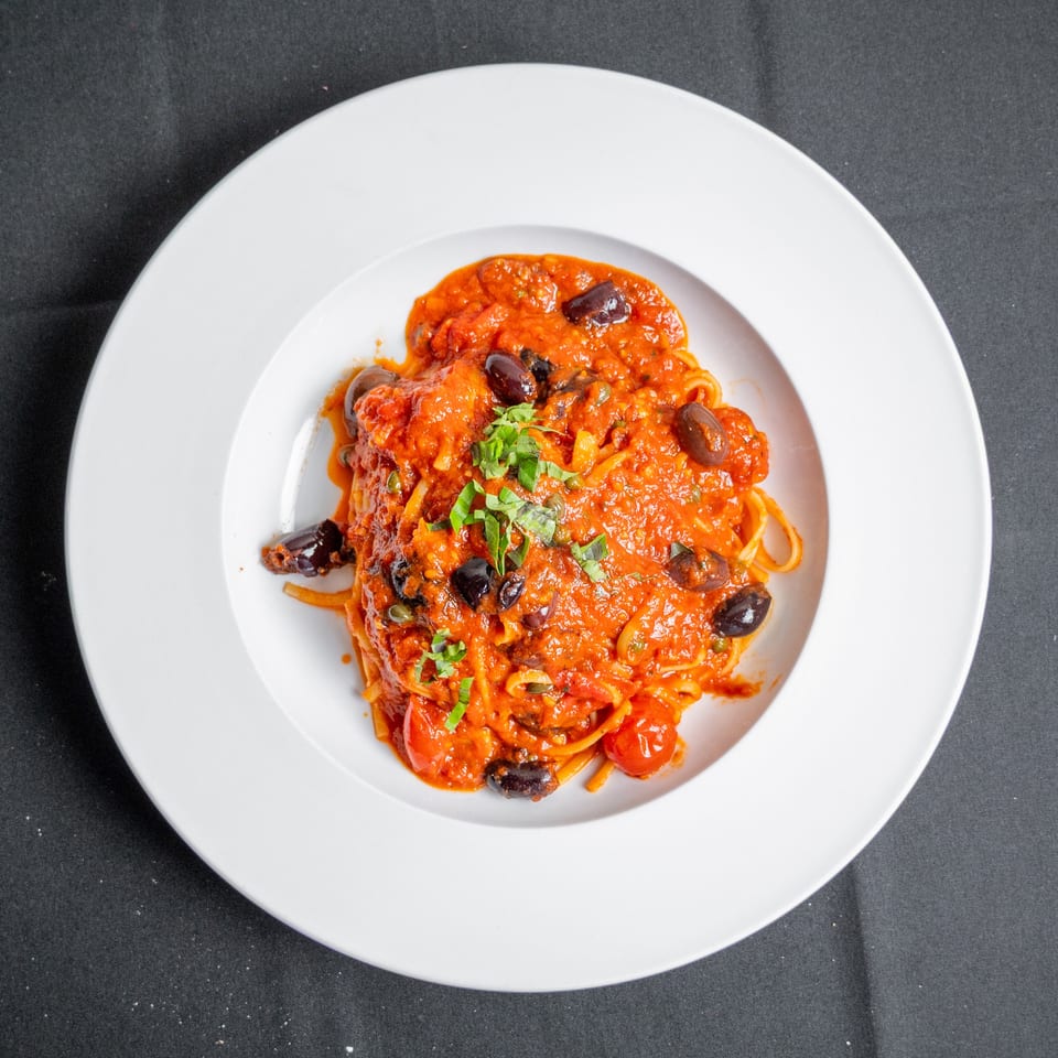 Spaghetti with red tomato sauce, black olives, cherry tomatoes, and fresh herbs in a white bowl.
