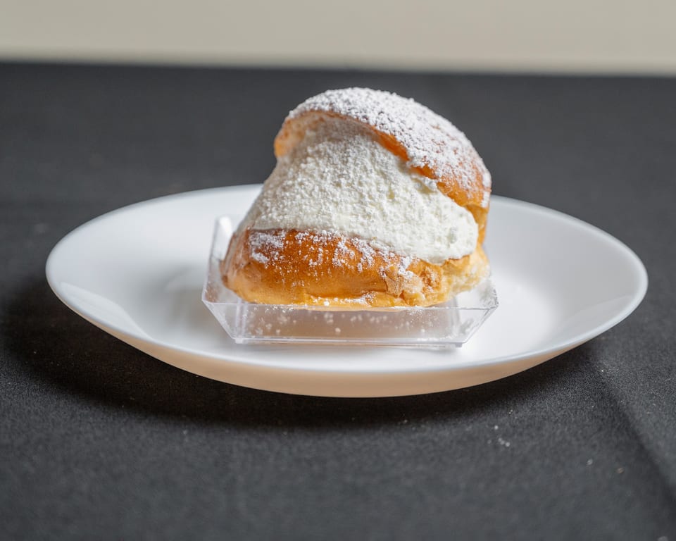 A golden-brown pastry bun filled with white cream and dusted with powdered sugar on a white plate.